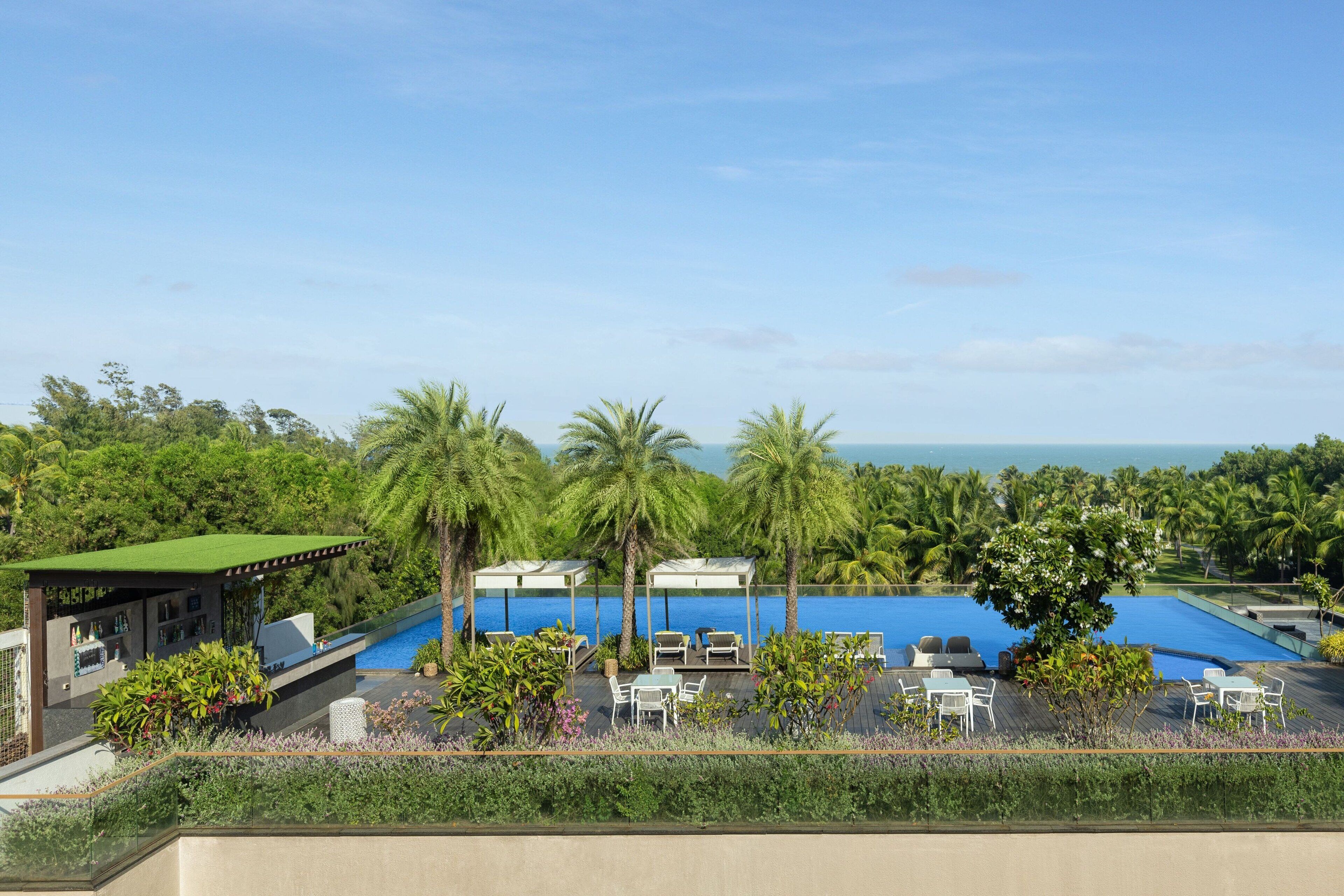 A rooftop pool area with lounge chairs and a bar, surrounded by palm trees and overlooking the ocean.