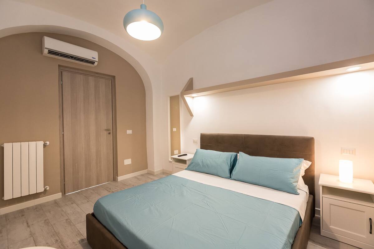 A bedroom with a bed, bedside table, nightstand, and an air conditioning unit.
