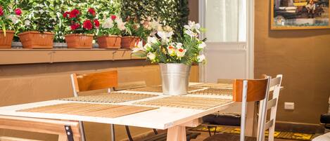 A dining area with a table set for four, surrounded by wooden chairs. There is a potted plant with white and red flowers in the center of the table.