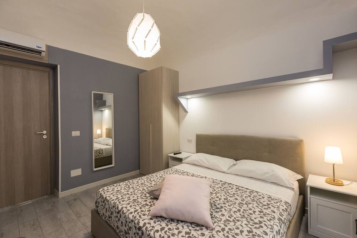 A bedroom with a bed, bedside tables, a mirror, and a hanging light fixture.