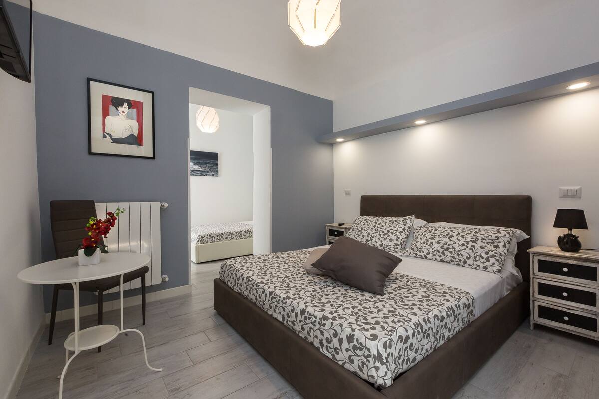 A modern bedroom with a large bed, bedside tables, a small table with a chair, and a framed picture on the wall.