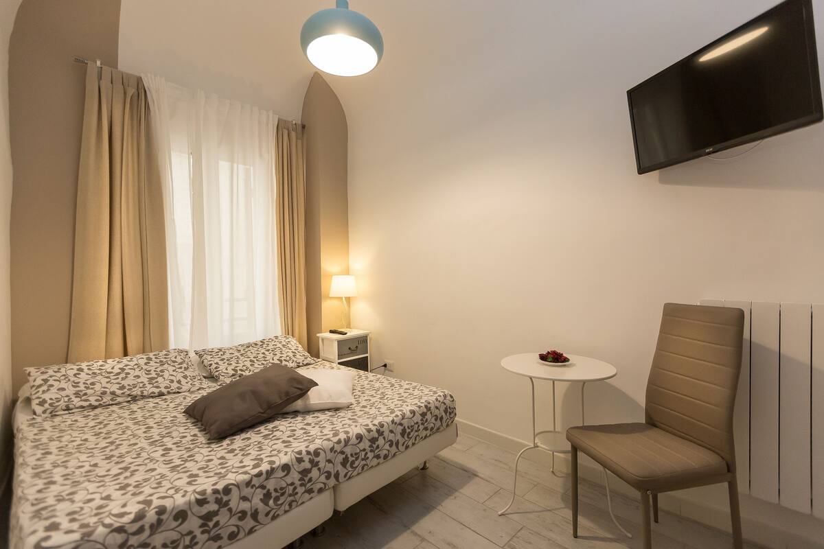 A hotel room with a bed, a chair, a TV, a small table with a lamp, and a window with curtains.