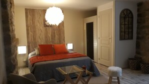 Un dormitorio con cama, una mesita de vidrio, una silla, un taburete pequeño, una lámpara colgante y una puerta.