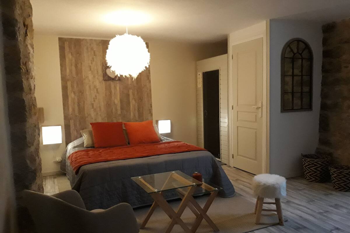 A bedroom with a bed, a glass table, a chair, a small stool, a hanging light fixture, and a door.
