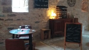 Un interior rústico con paredes de piedra, una mesa redonda con sillas, un taburete pequeño, una pizarra con escritura y un reloj en la pared.