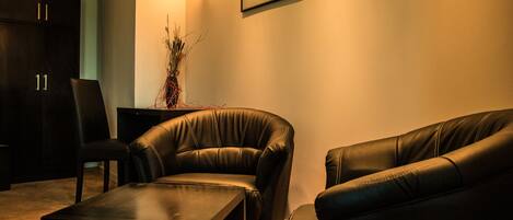 A dark leather sofa, a matching armchair, and a small table in a dimly lit room with a wall-mounted light and a framed picture.