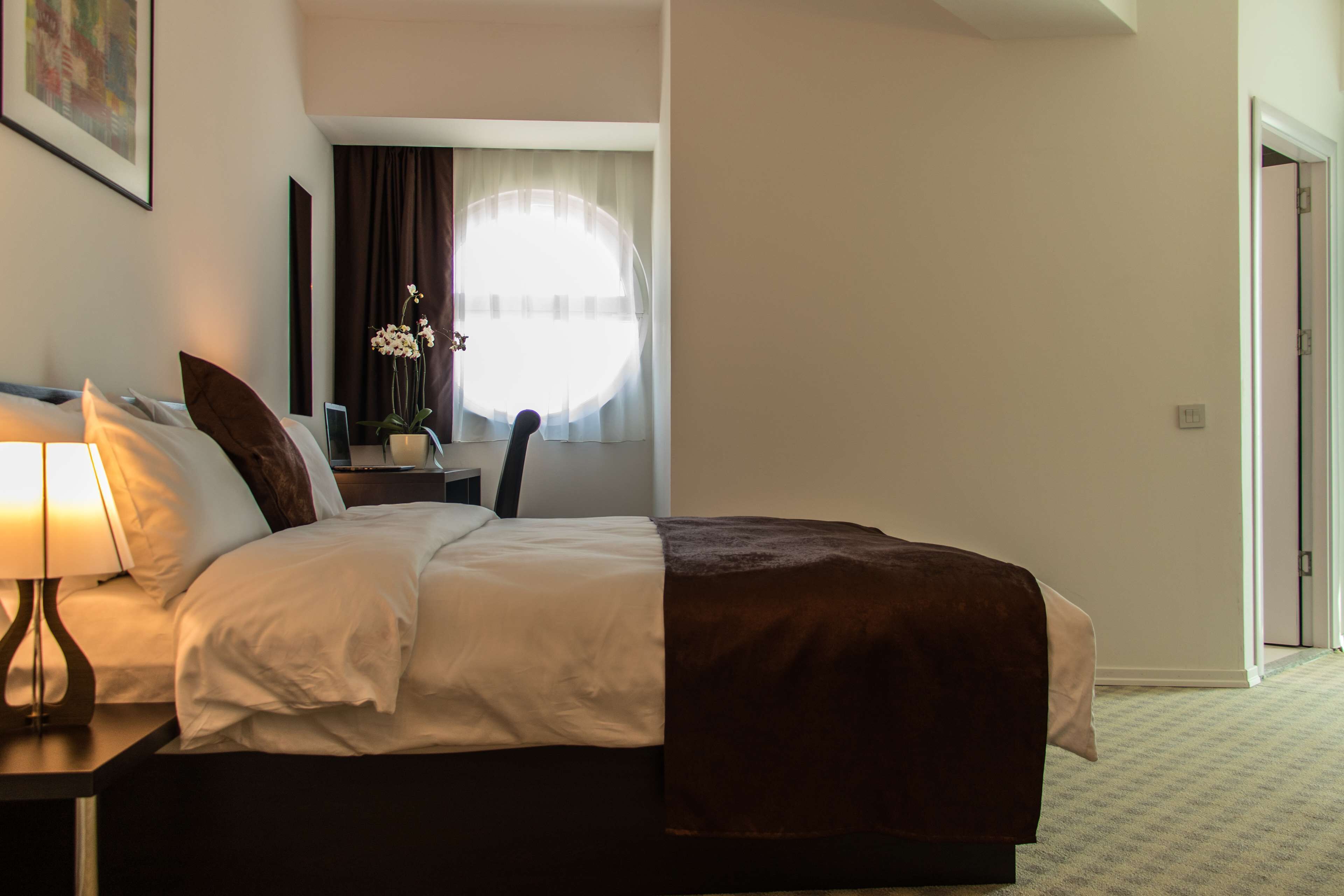 Une chambre d’hôtel comprenant un lit, une table de chevet, une lampe, un bureau avec une chaise, une fenêtre avec des rideaux et une porte.