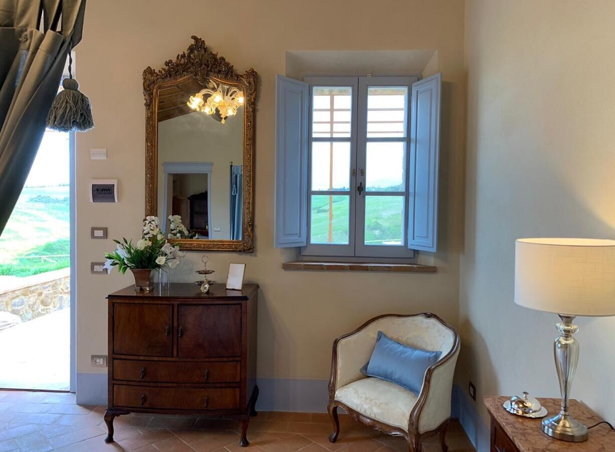 A room with a wooden dresser, a chair, a mirror, a window with shutters, and a lamp.