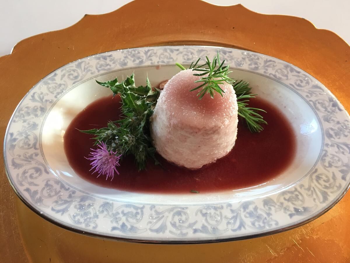 Un plato con un alimento, posiblemente un buñuelo, adornado con hierbas y una flor morada, servido en un plato estampado.