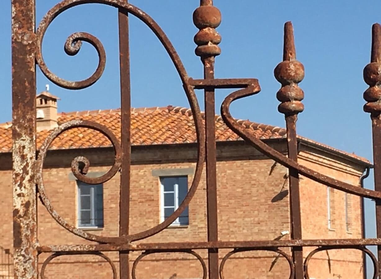A rustic metal gate with a spiral design, a brick building with a tiled roof, and a window in the background.