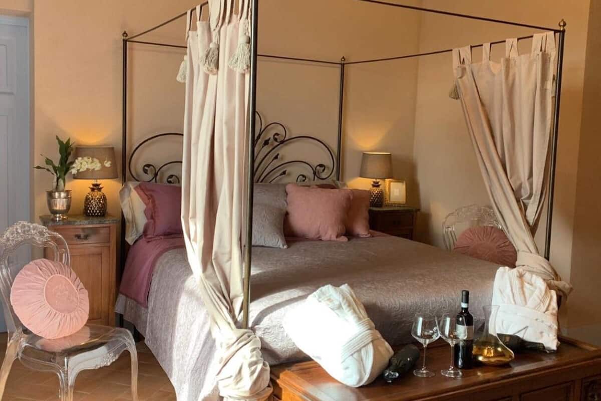 A four-poster canopy bed with a beige bedspread, two bedside tables with lamps, a clear chair with a pink cushion, and a wooden nightstand with a potted plant.