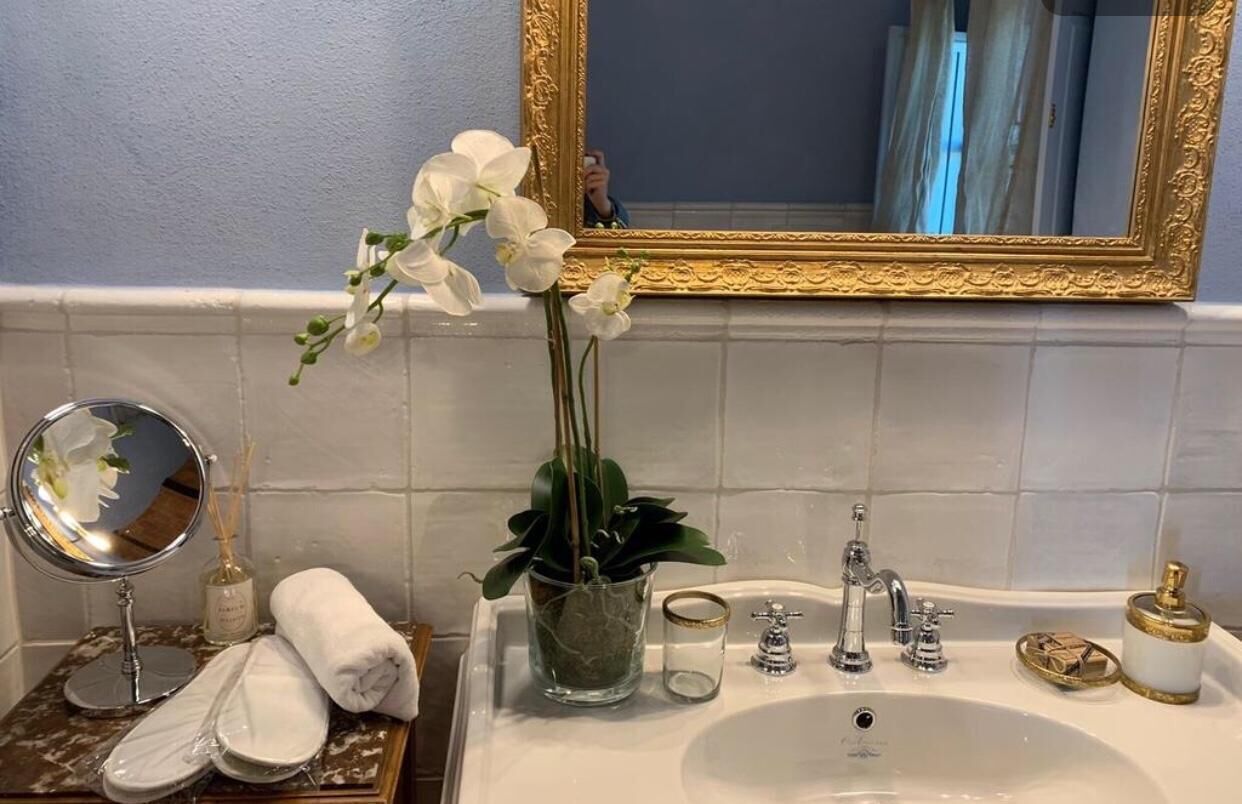 Une salle de bain avec un lavabo blanc, un miroir encadré d’or, une orchidée en pot, un petit récipient en verre et une serviette.