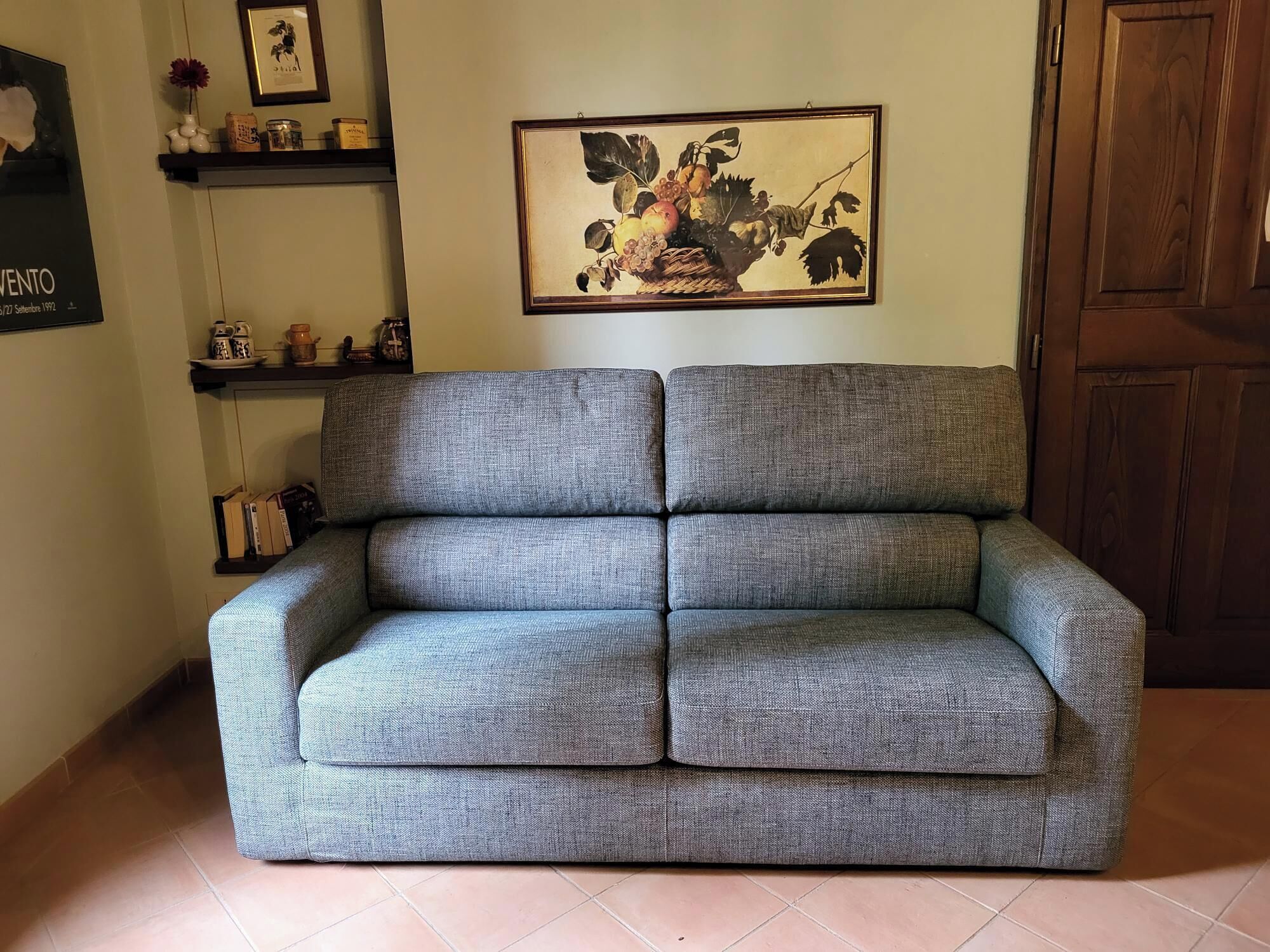 A grey sofa in a room with a wooden door, a framed artwork on the wall, and shelves with various items.