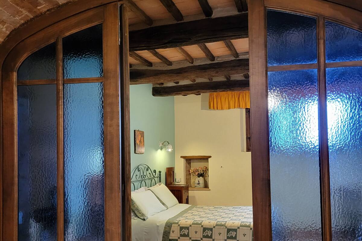 Una habitación con cama, mesita de noche, un cuadro enmarco y una ventana con vidrio esmerilado.