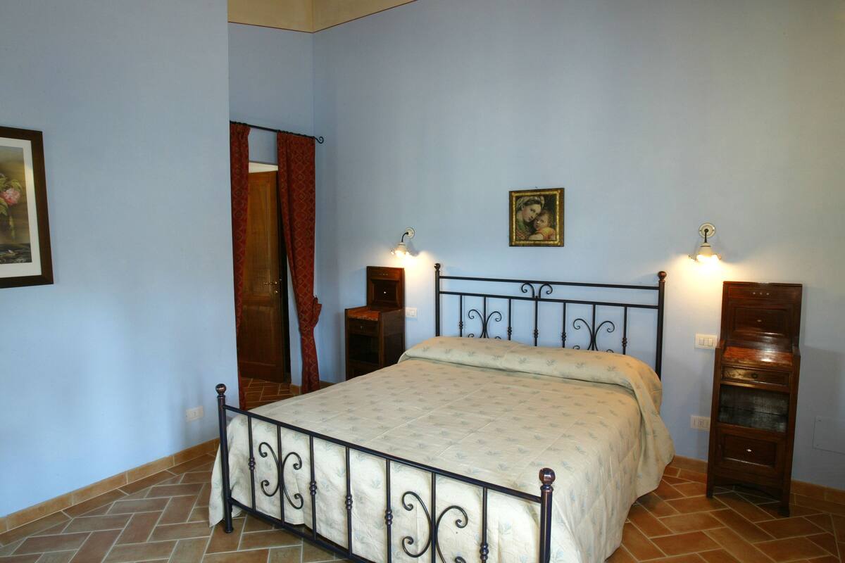 Un dormitorio con cama, mesita de noche, cómoda, un cuadro y una puerta con cortina.