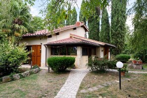 Una casa con tejado de baldosas, un camino de piedra que lleva a la entrada, y un jardín con diversas plantas y árboles.