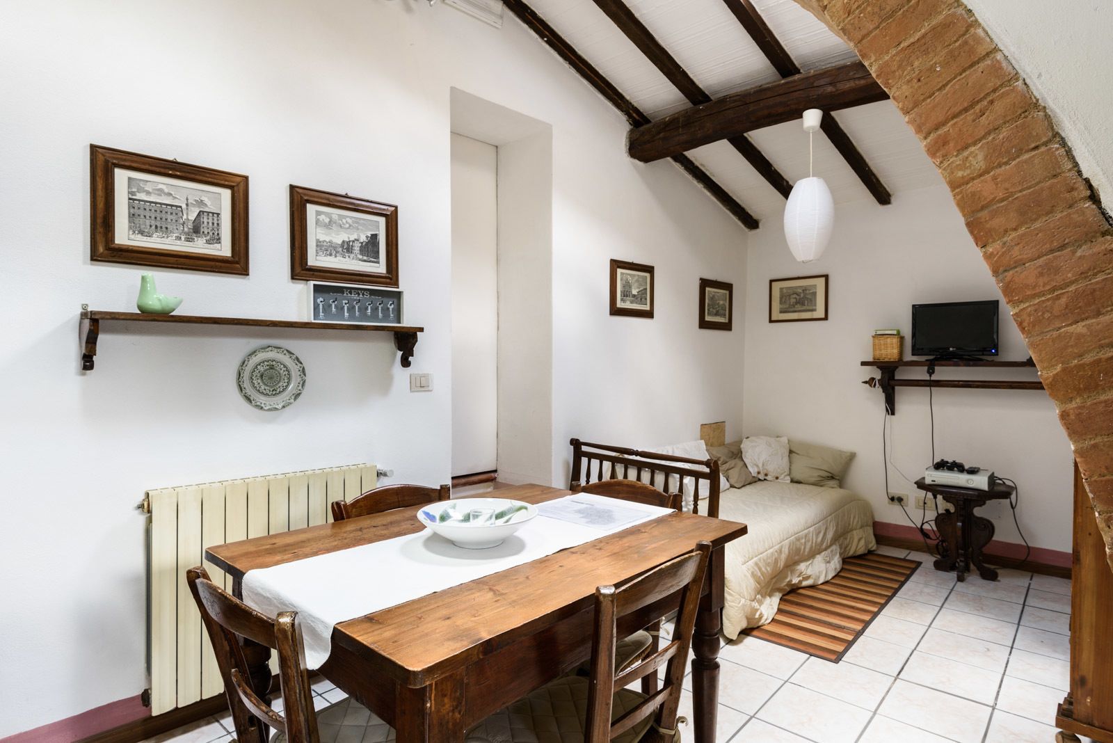 A cozy dining area with a wooden table and chairs, a radiator, and a bed with a white comforter in the background.