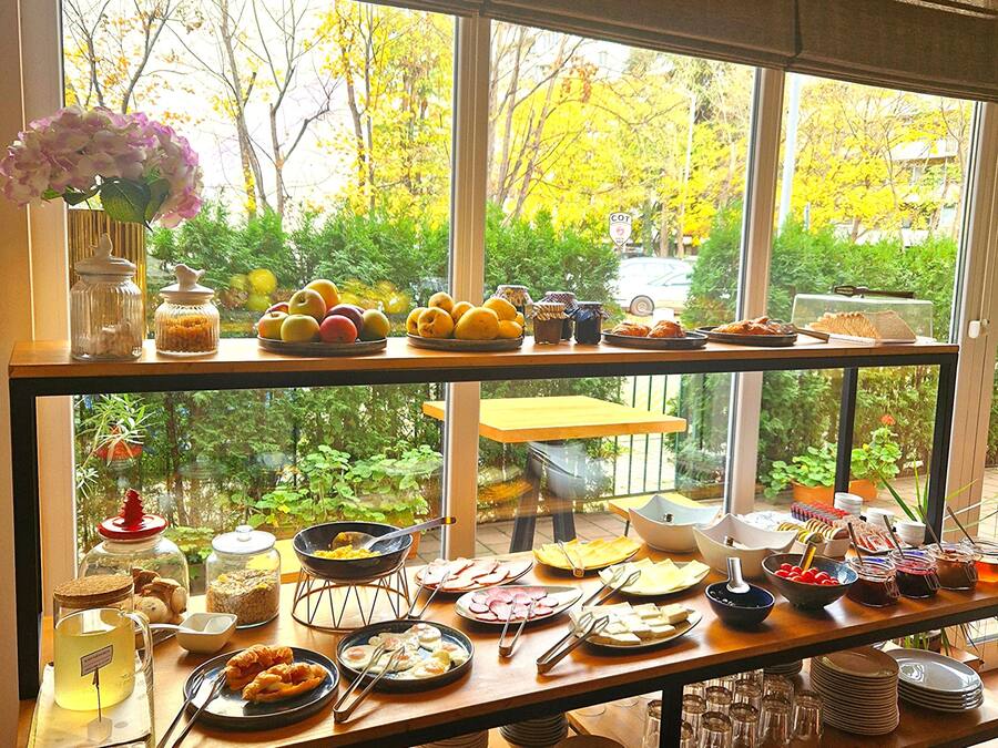 Un bufé de desayuno con diversas opciones de comida y utensilios, con vista a un paisaje de árboles y un automóvil a través de una ventana.