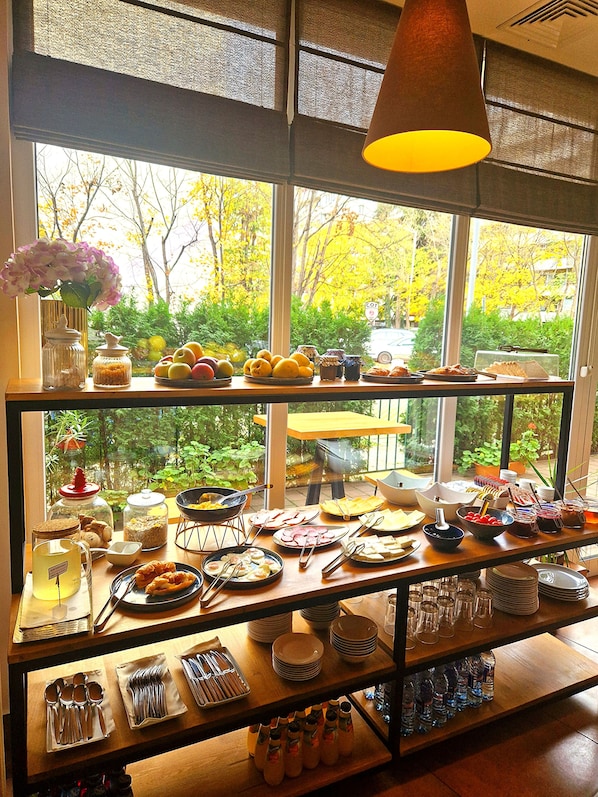Un bufé de desayuno con diversas opciones de comida y utensilios, con vista a un paisaje de árboles y un automóvil a través de una ventana.