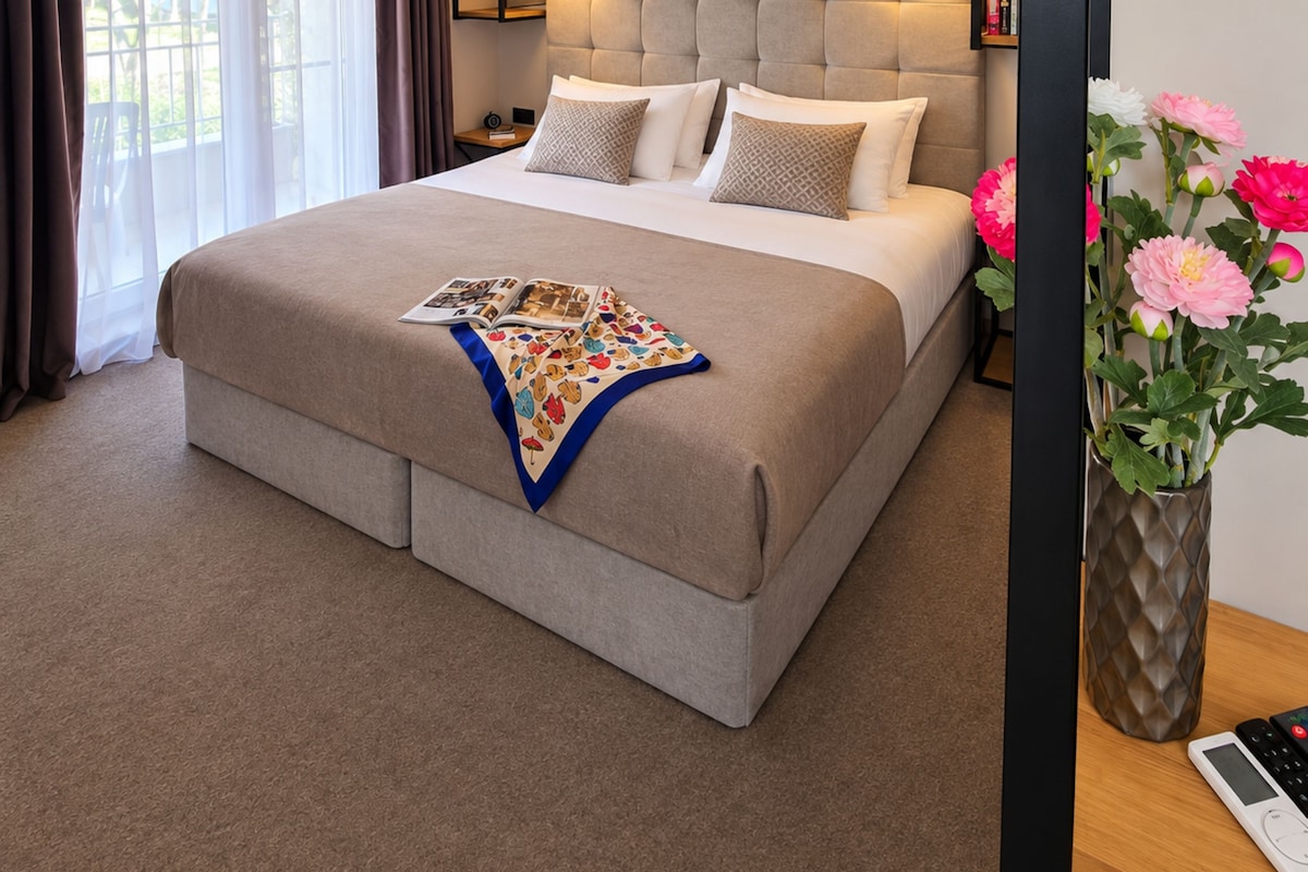 Quarto de hotel com uma cama grande, cabeceira com iluminação integrada, mesa de cabeceira com abajur e um vaso com flores.