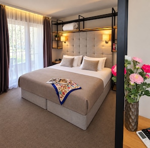 Quarto de hotel com uma cama grande, cabeceira com iluminação integrada, mesa de cabeceira com abajur e um vaso com flores.