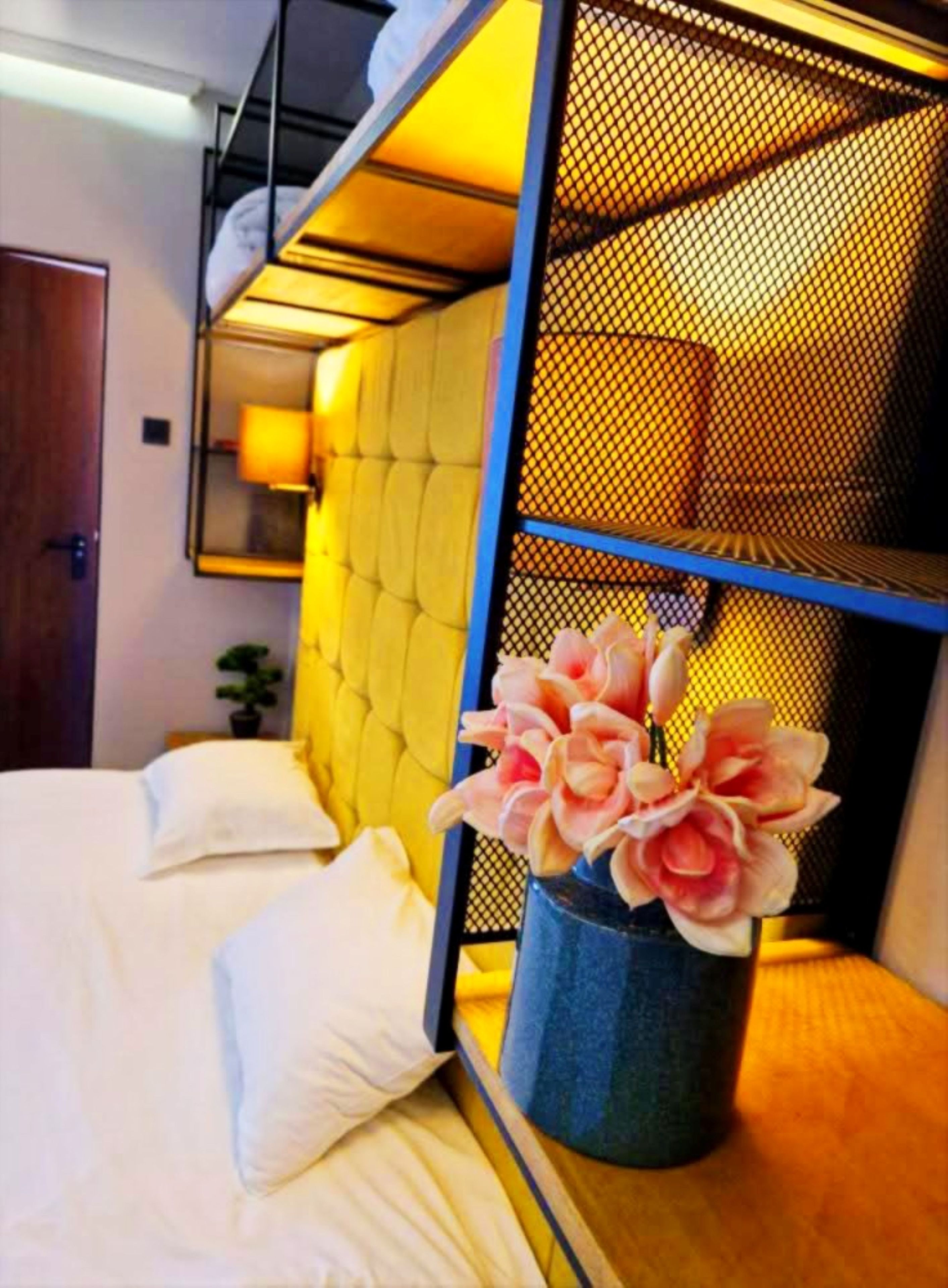 Una cama individual con cabecera amarilla, una mesita de noche con un florero y un espejo en la pared.