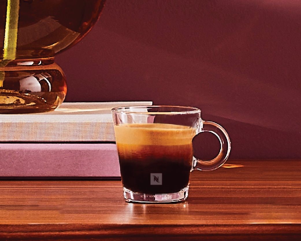 A clear glass cup with a handle on a wooden surface, a book, and a decorative object in the background.