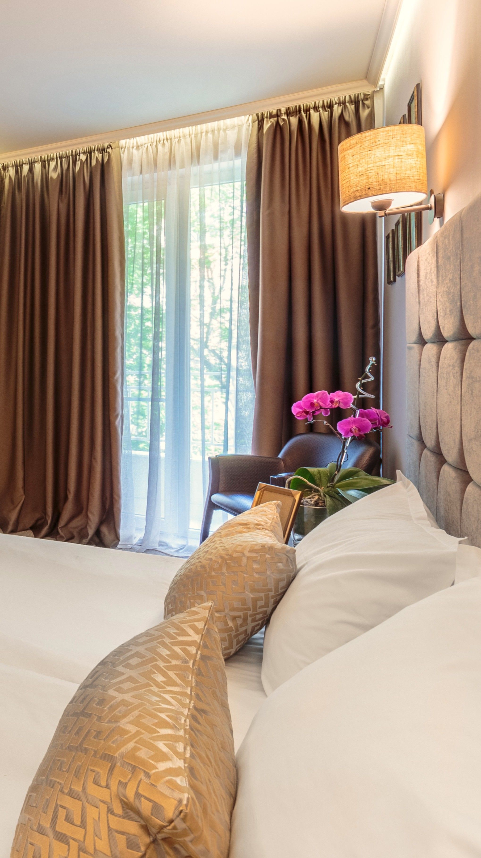 Une chambre d’hôtel avec un lit, des rideaux, une lampe, une chaise et un vase avec des fleurs.