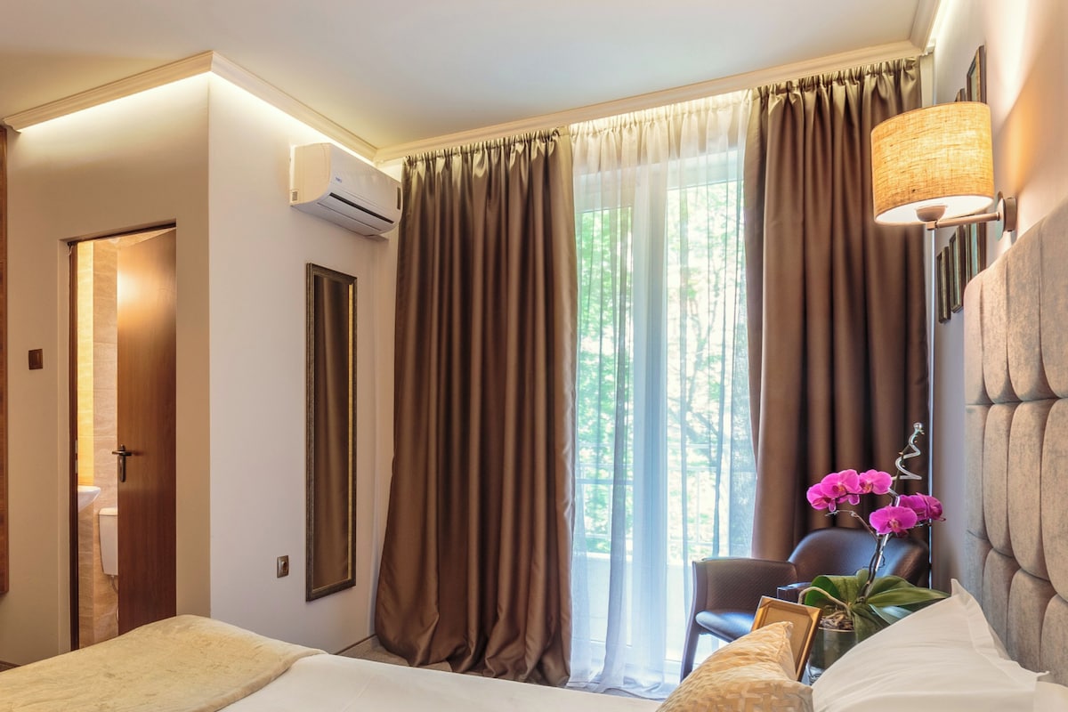 Quarto de hotel com uma cama, uma cadeira, um abajur e uma janela com cortinas.