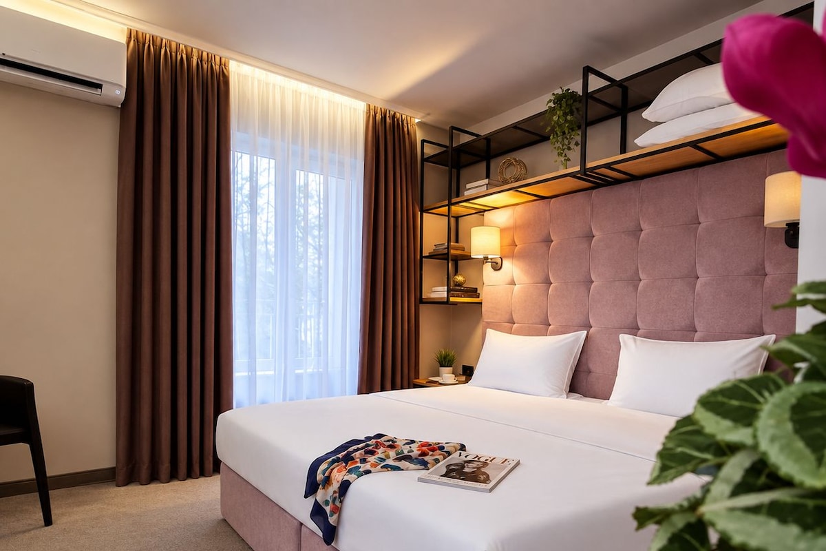 Quarto de hotel com uma cama grande, uma mesa de cabeceira com abajur, uma prateleira na parede com objetos decorativos e uma janela com cortinas.