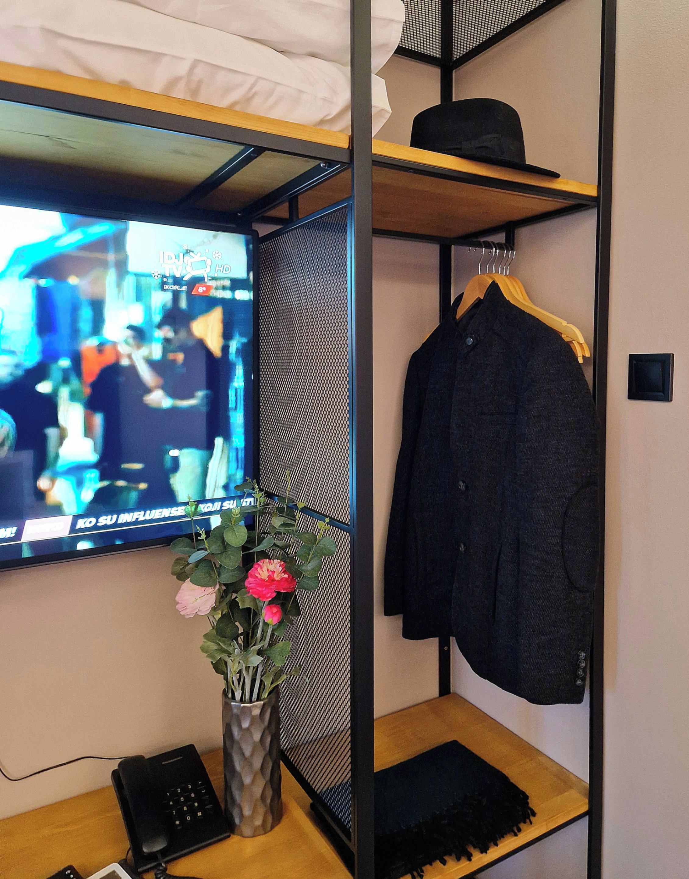 Una habitación con una televisión montada en la pared, un escritorio con un teclado y un teléfono, una estantería con una chaqueta y una planta con flores.