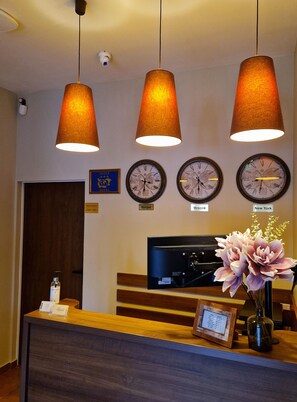 Área de recepção de hotel com três relógios mostrando diferentes fusos horários, balcão de atendimento com um computador e um vaso de flores, e uma placa indicando que o acesso é exclusivo para funcionários.