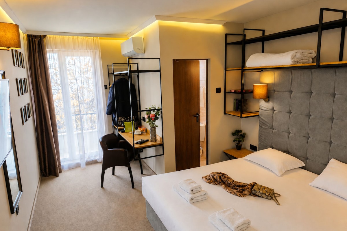 Quarto de hotel com cama, escrivaninha, cadeira, guarda-roupa e janela com cortinas.