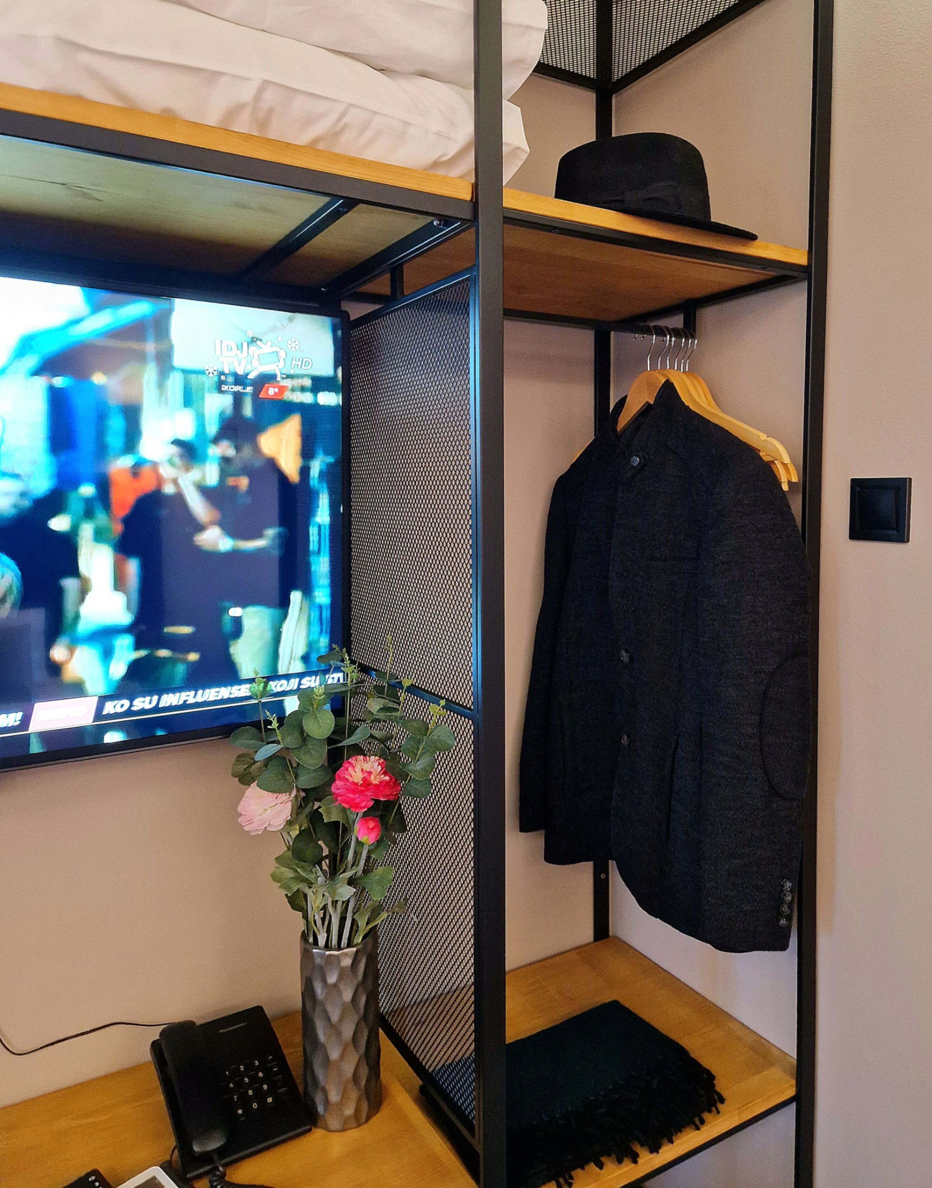 Ein Zimmer mit einem an der Wand befestigten Fernseher, einem Schreibtisch mit Tastatur und Telefon sowie einem Regal mit einem Mantel und einer Vase mit Blumen.