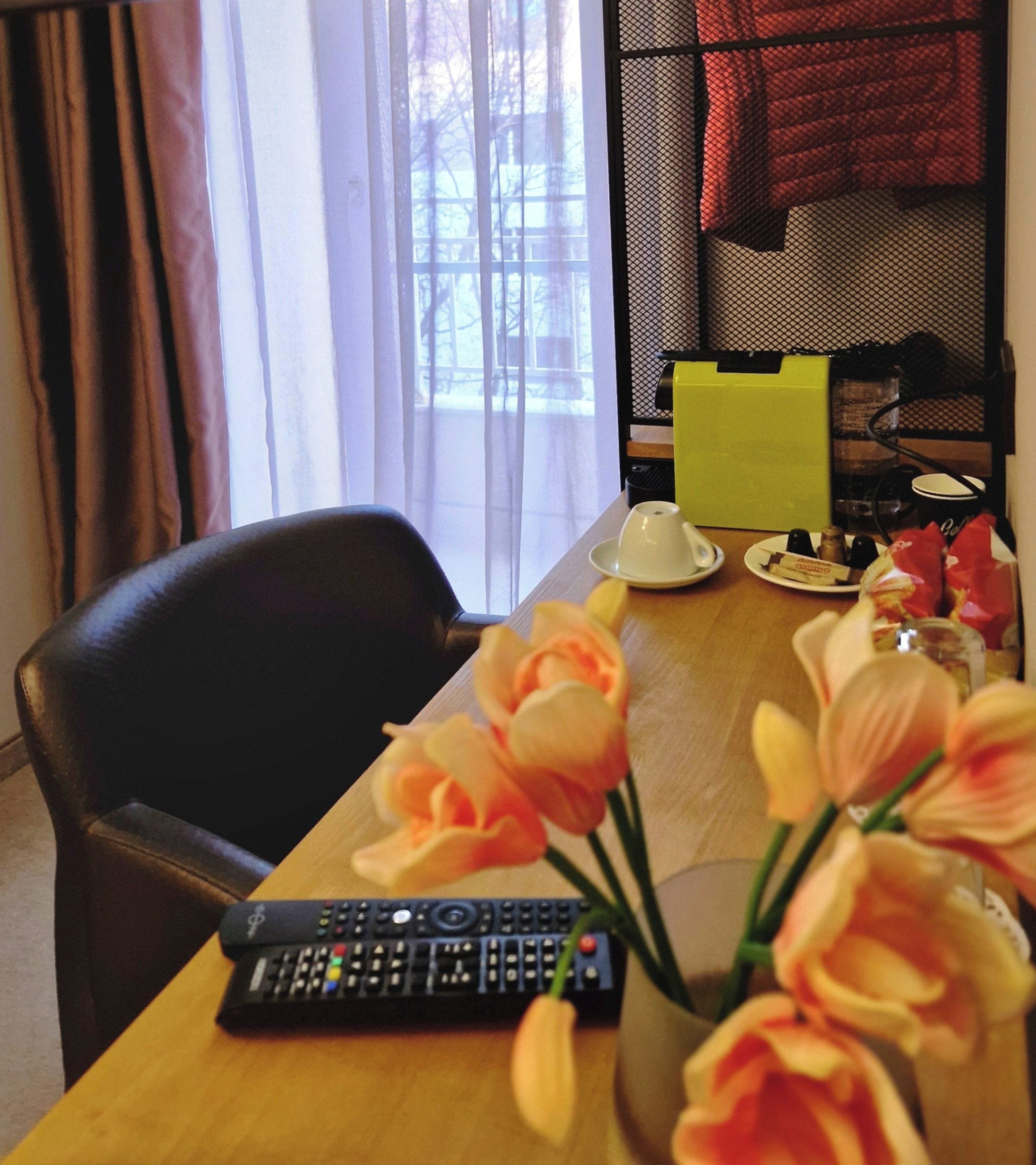 Habitación de hotel con una mesa de madera, un control remoto, un florero con flores y una ventana con cortinas.
