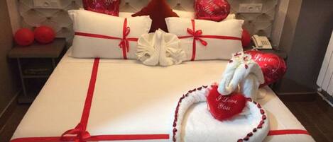 A neatly made bed with white linens and red ribbons, adorned with heart-shaped pillows and a decorative pillow with a heart and the text "I Love You".