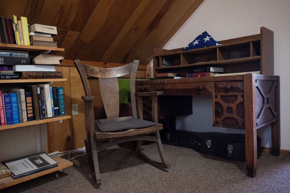 Une chaise en bois dans une pièce où se trouvent des étagères à livres, un bureau et une housse de guitare.