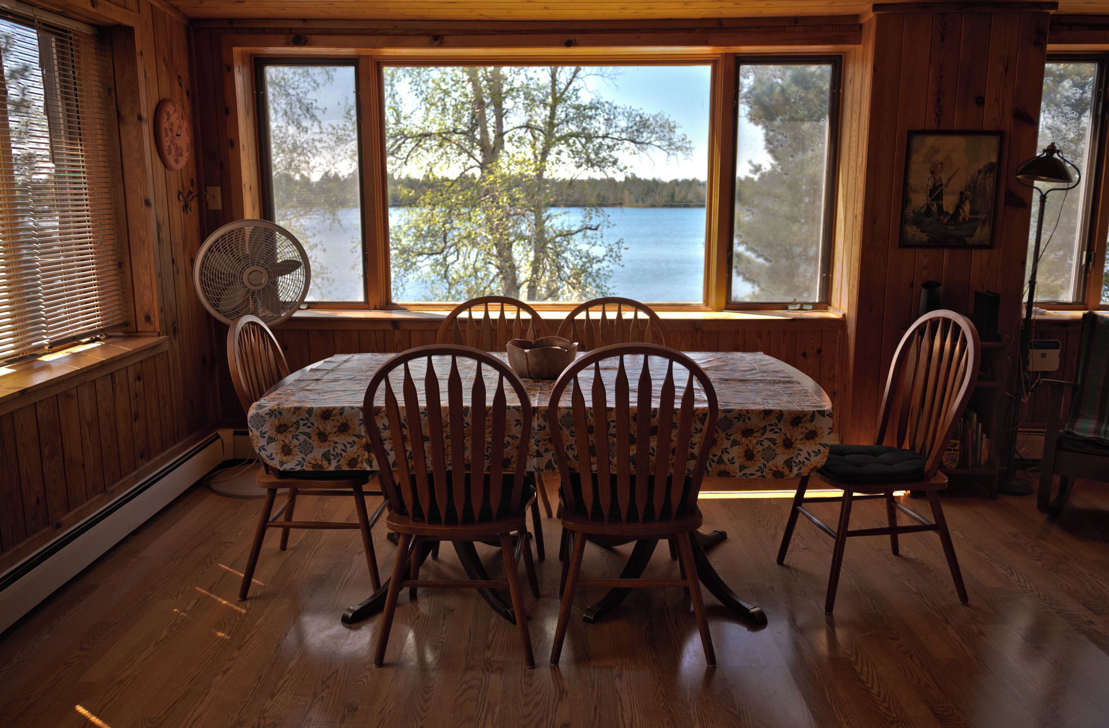 Une salle à manger en bois avec une table et des chaises, de grandes fenêtres donnant sur un lac et un ventilateur au sol.