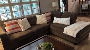Ein Wohnzimmer mit einem braunen Ecksofa, einem Couchtisch mit einer Pflanze und einem Fenster mit Jalousien.