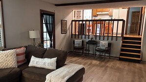 Ein gemütliches Wohnzimmer mit einem braunen Sofa, einer weißen Decke und einem Kissen mit der Aufschrift 'ALL ARE WELCOME HERE'. Eine Treppe führt zu einer Küchenzeile mit Holzschränken und einem Kühlschrank.