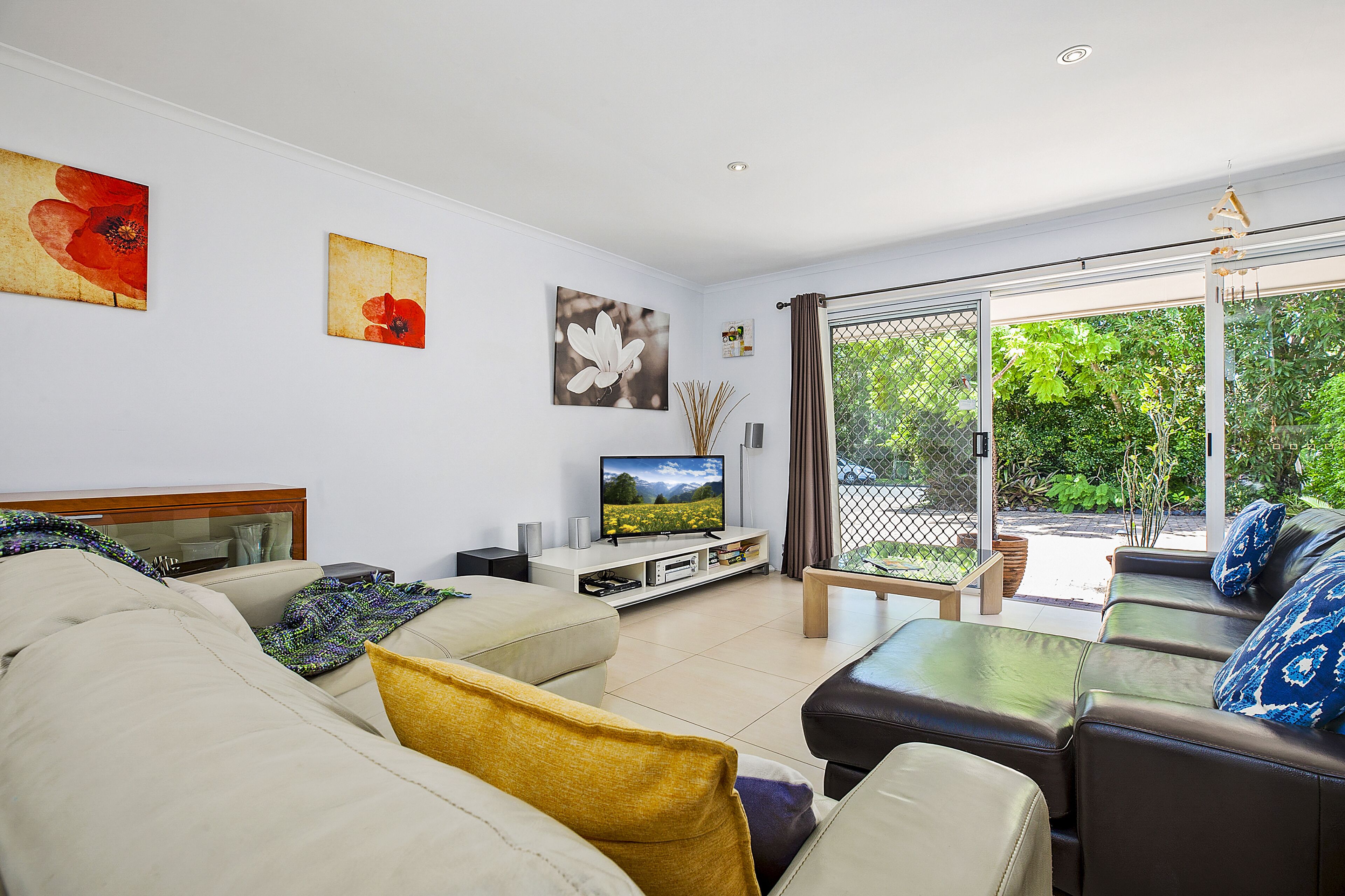 A living room with a sofa, coffee table, and TV stand. There are two paintings on the wall and a sliding glass door leading to an outdoor area.
