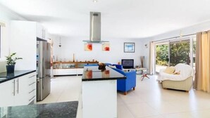 A modern kitchen with a central island, a dining area with a blue chair, and a sofa. - 'No-Steps Noosa' - Home Away From Home 🏠  (Noosa Heads)