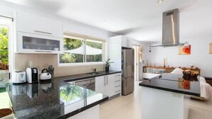 A modern kitchen with white cabinets, a black countertop, a stainless steel refrigerator, and a window with a view of greenery. - 'No-Steps Noosa' - Home Away From Home 🏠  (Noosa Heads)