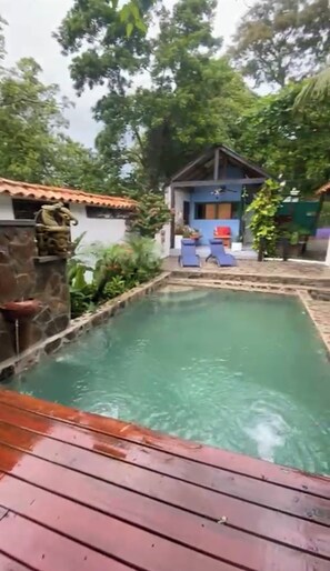 Une piscine avec une terrasse en bois au premier plan, entourée d’une végétation luxuriante et d’un petit bâtiment doté d’un toit bleu.