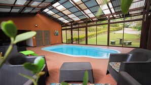 Una piscina interior con vistas a un jardín ajardinado, rodeada de mobiliario de mimbre y un techo cubierto.