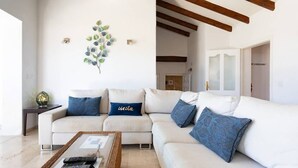 Un salón con sofá blanco, cojines azules, una mesita de centro con mandos a distancia y un adorno en la pared.