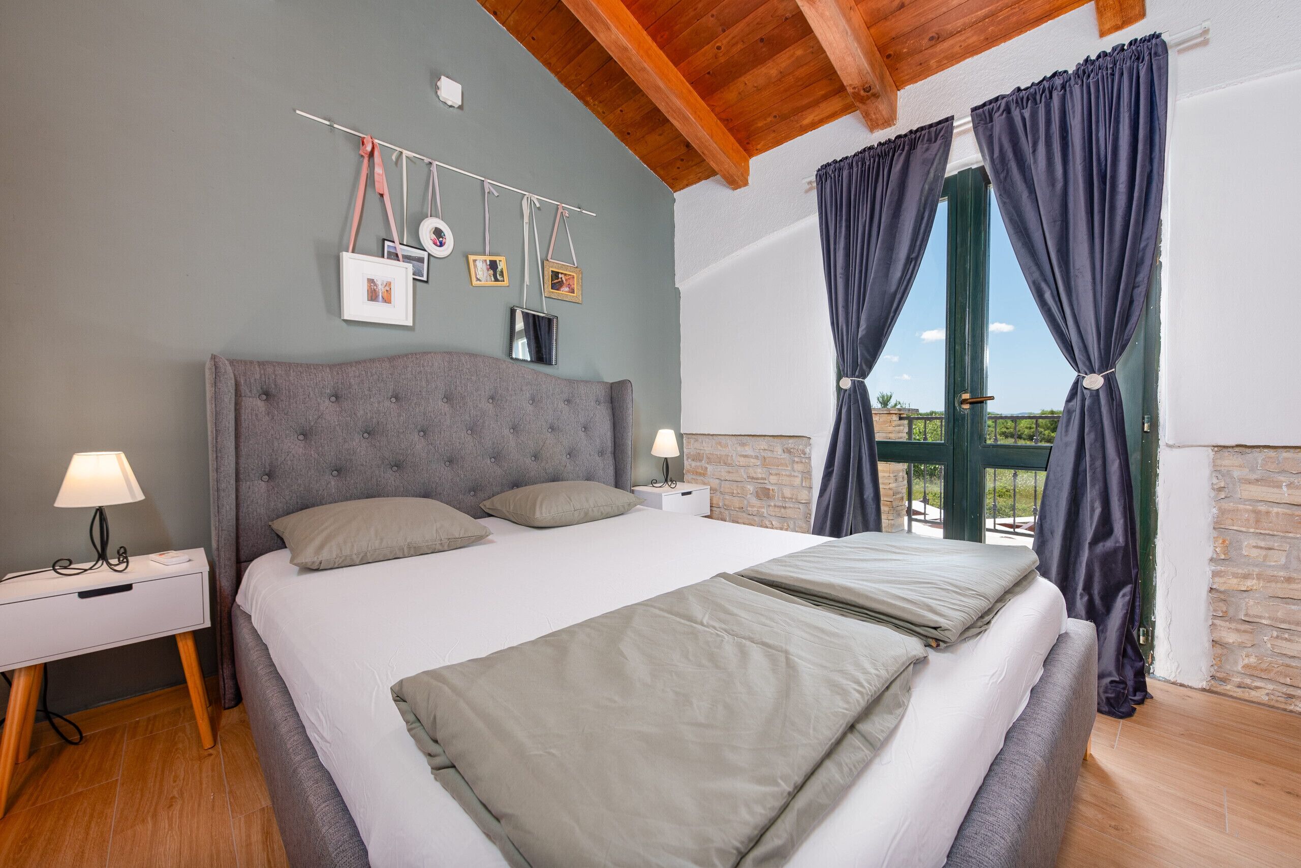 Un dormitorio con una cama grande, una mesita de noche, una lámpara y vistas al exterior a través de una ventana con cortinas.