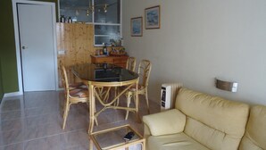 Un coin repas avec une table en verre et des chaises en osier, un canapé beige et une cuisine dotée de meubles en bois et d’une fenêtre.