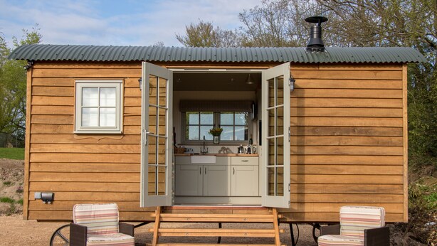 Eine Holzhütte mit offener Tür, aus der eine Küche, zwei Korbstühle und ein kleiner Holztisch sichtbar sind.