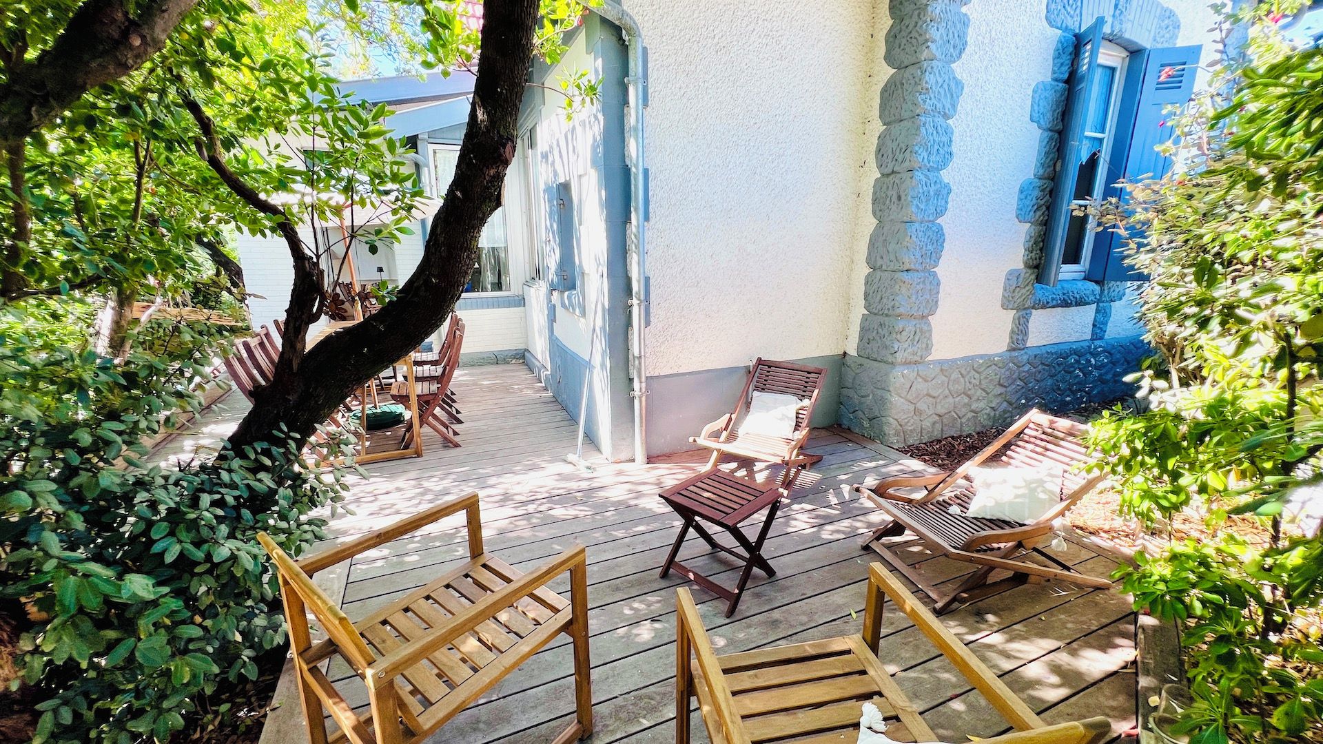 Eine Holzterrasse mit Gartenmöbeln, einem Baum und einem Gebäude mit blauen Fensterläden.